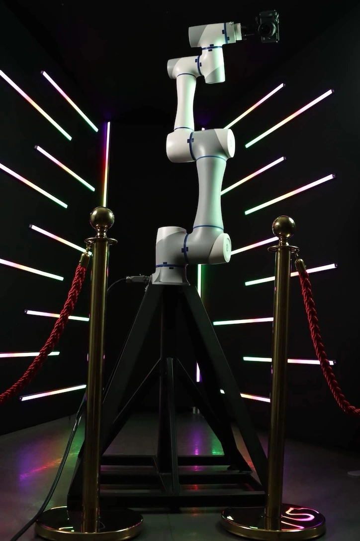 A high-tech white robotic camera arm mounted on a tripod for interactive event videography, set against a futuristic LED neon light tube background.